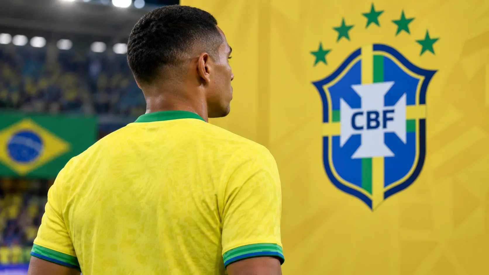 Jogador com camisa amarela da Seleção visto de costas em estádio, diante de painel amarelo com escudo da CBF e bandeira do Brasil ao fundo.