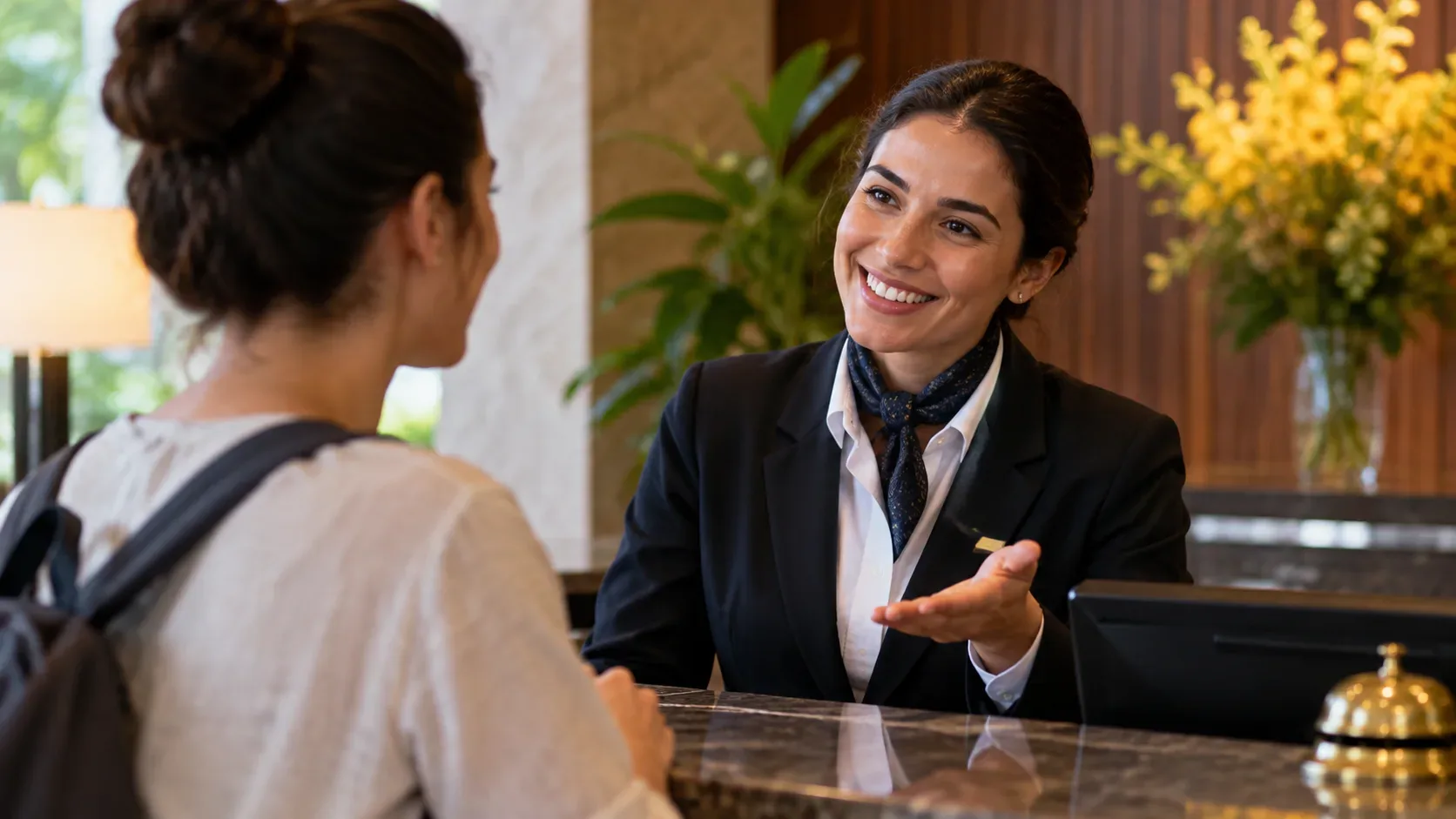 Atendente de hotel sorridente conversa com uma hóspede e faz um gesto acolhedor diante do balcão da recepção.