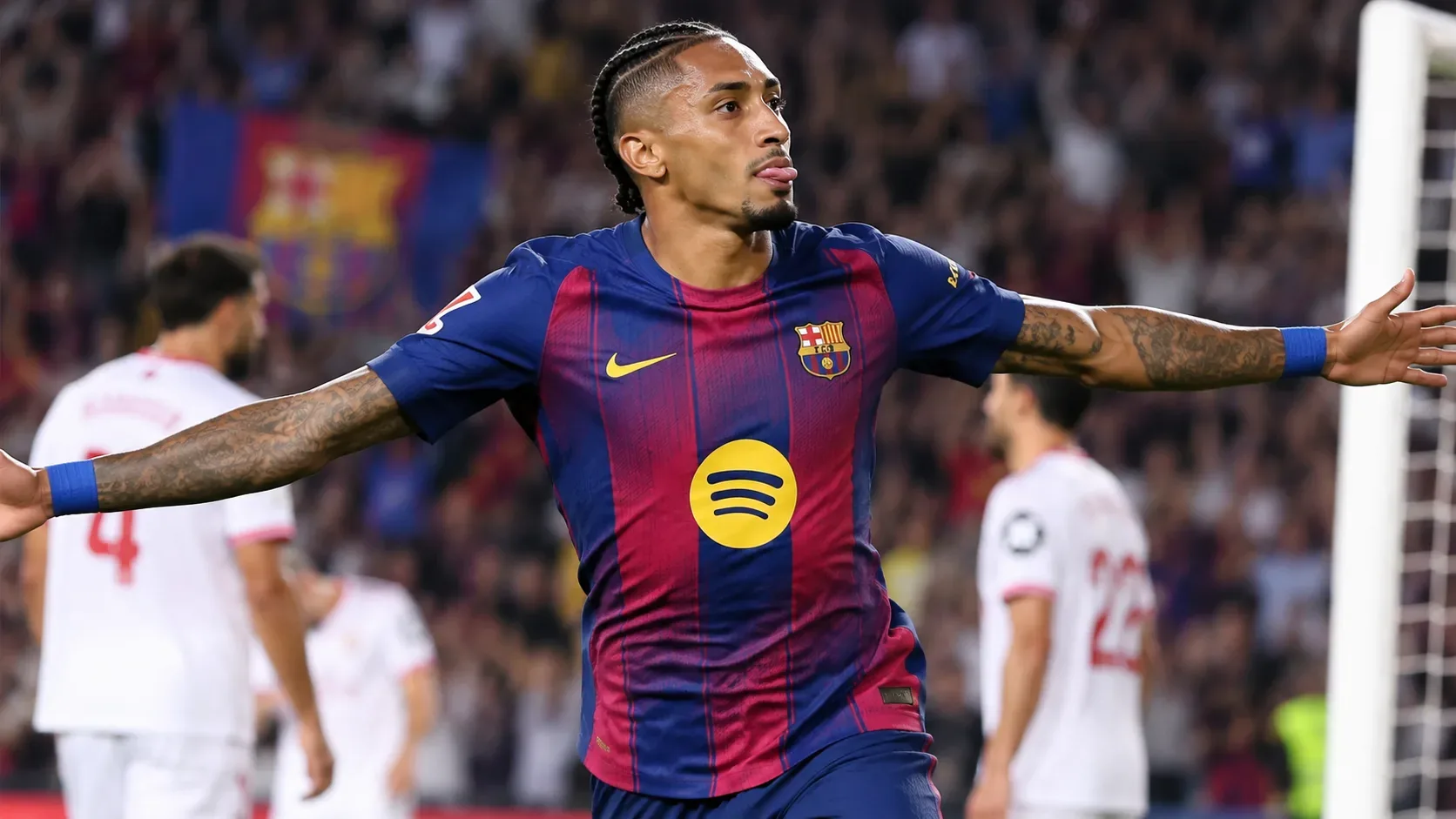 Raphinha comemora com os braços abertos em campo, com jogadores do Sevilla ao fundo diante do gol.