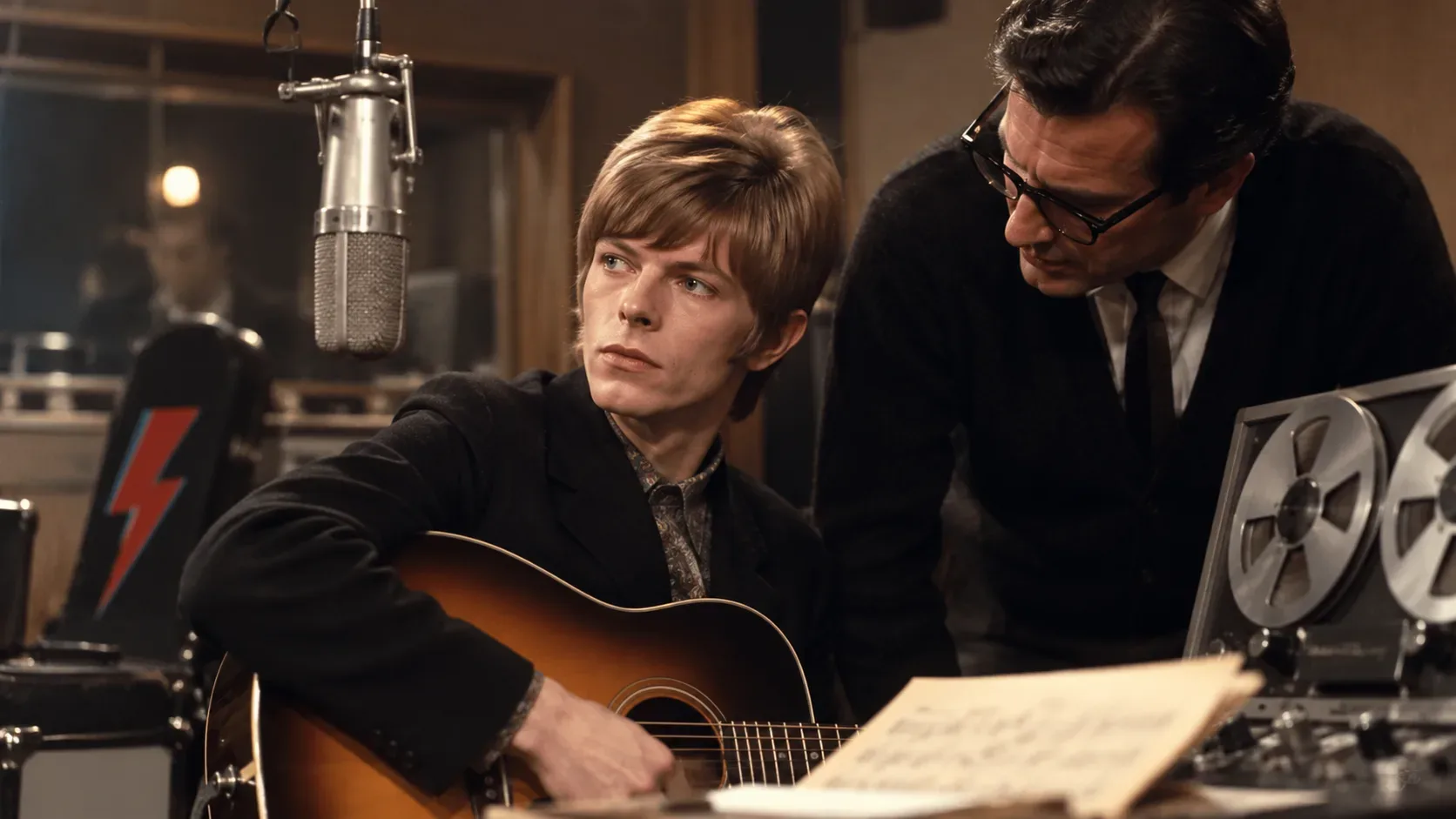 Representação de um jovem David Bowie com violão ao lado de um produtor em estúdio dos anos 1960, com microfone suspenso e gravador de rolo.