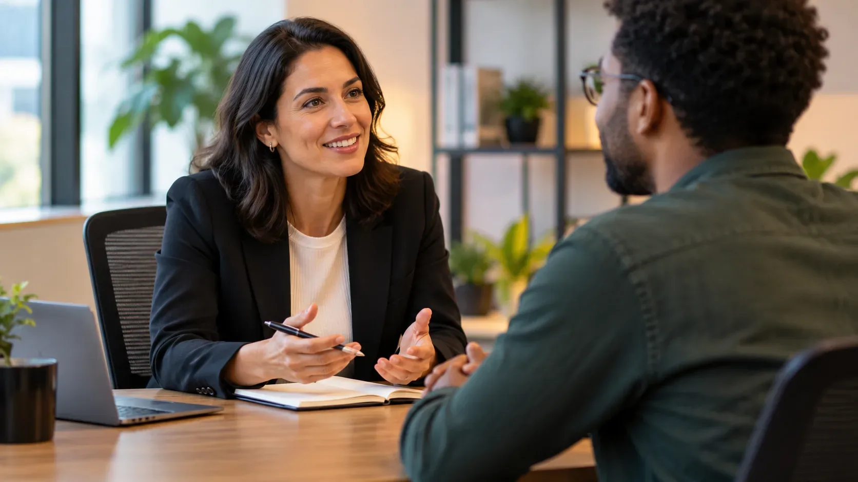 Gestora conversa com colaborador em uma reunião individual, com notebook e caderno sobre a mesa em um escritório iluminado.
