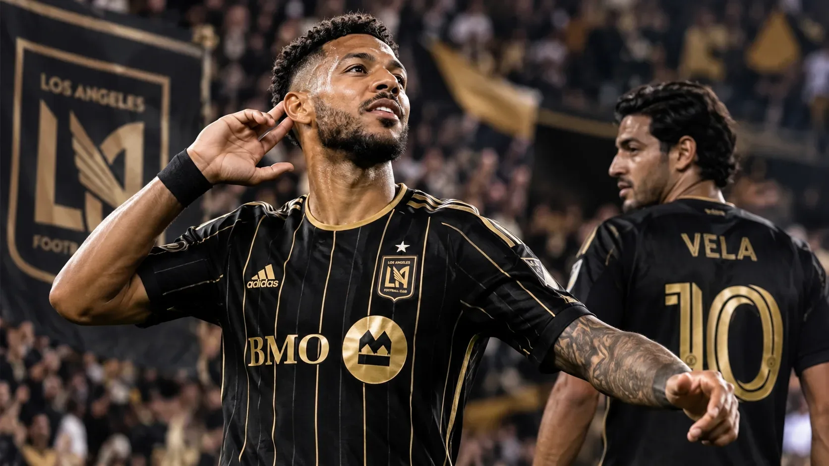 Atacante do LAFC comemora em primeiro plano, com outro jogador do clube ao fundo em um estádio com torcida nas cores preto e dourado.
