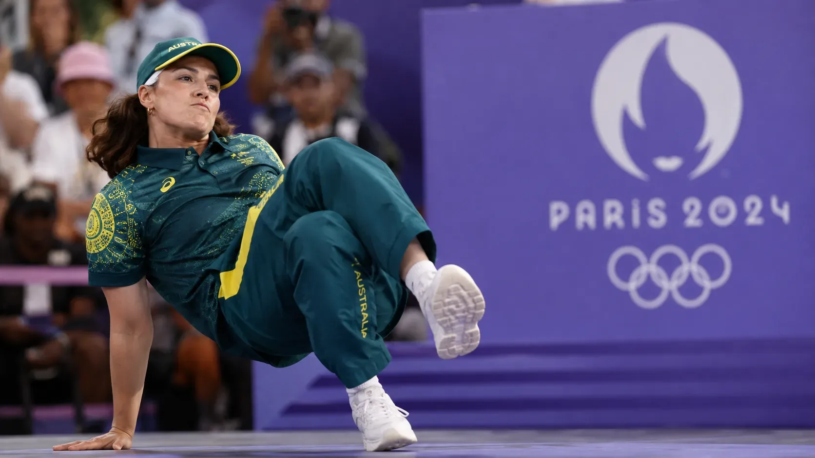 Competidora australiana de breaking em movimento no chão de uma arena olímpica, com uniforme verde e amarelo e fundo roxo de Paris 2024.