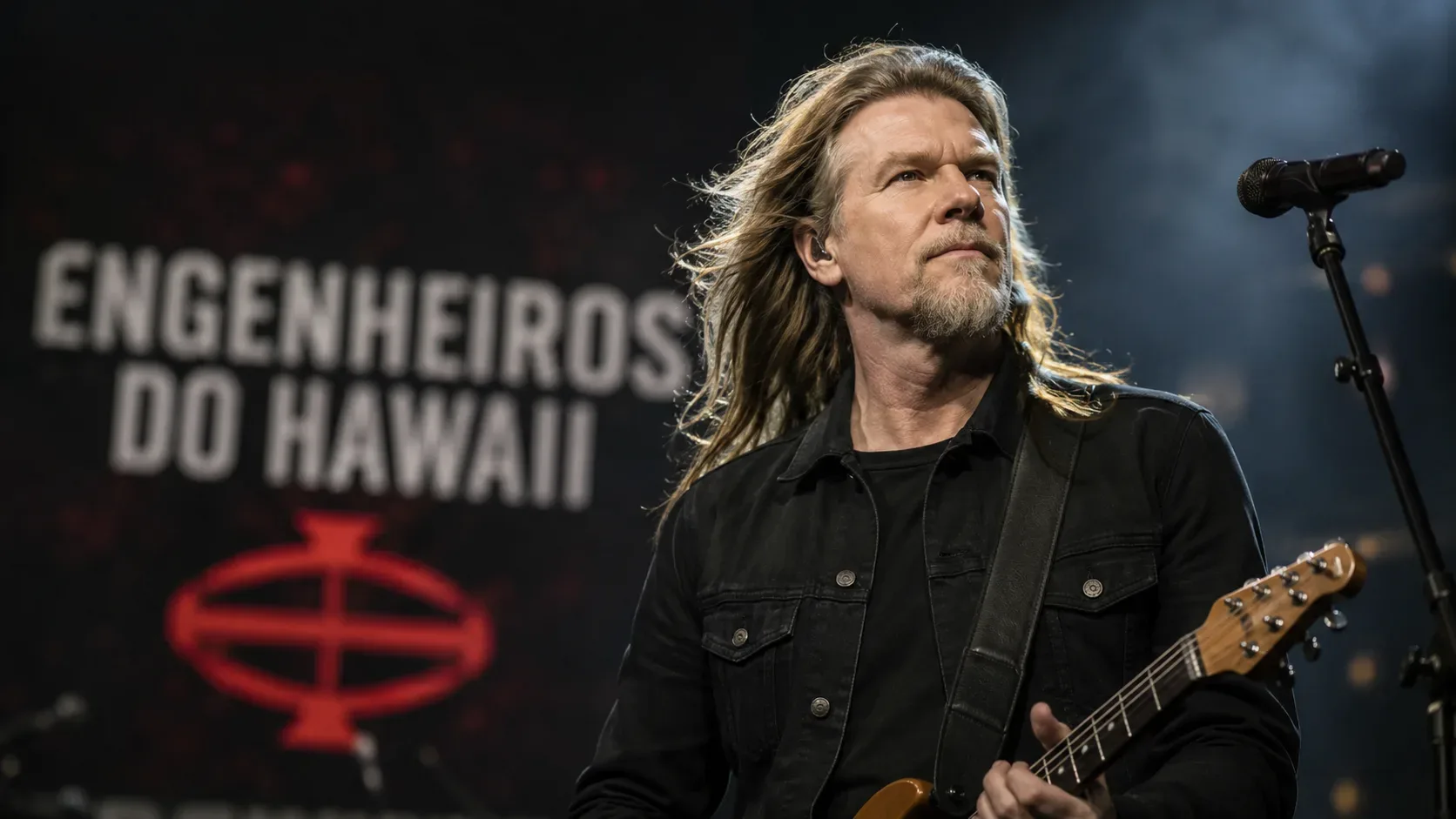 Músico de rock de cabelos longos tocando guitarra em palco escuro, com microfone e o nome Engenheiros do Hawaii desfocado ao fundo.