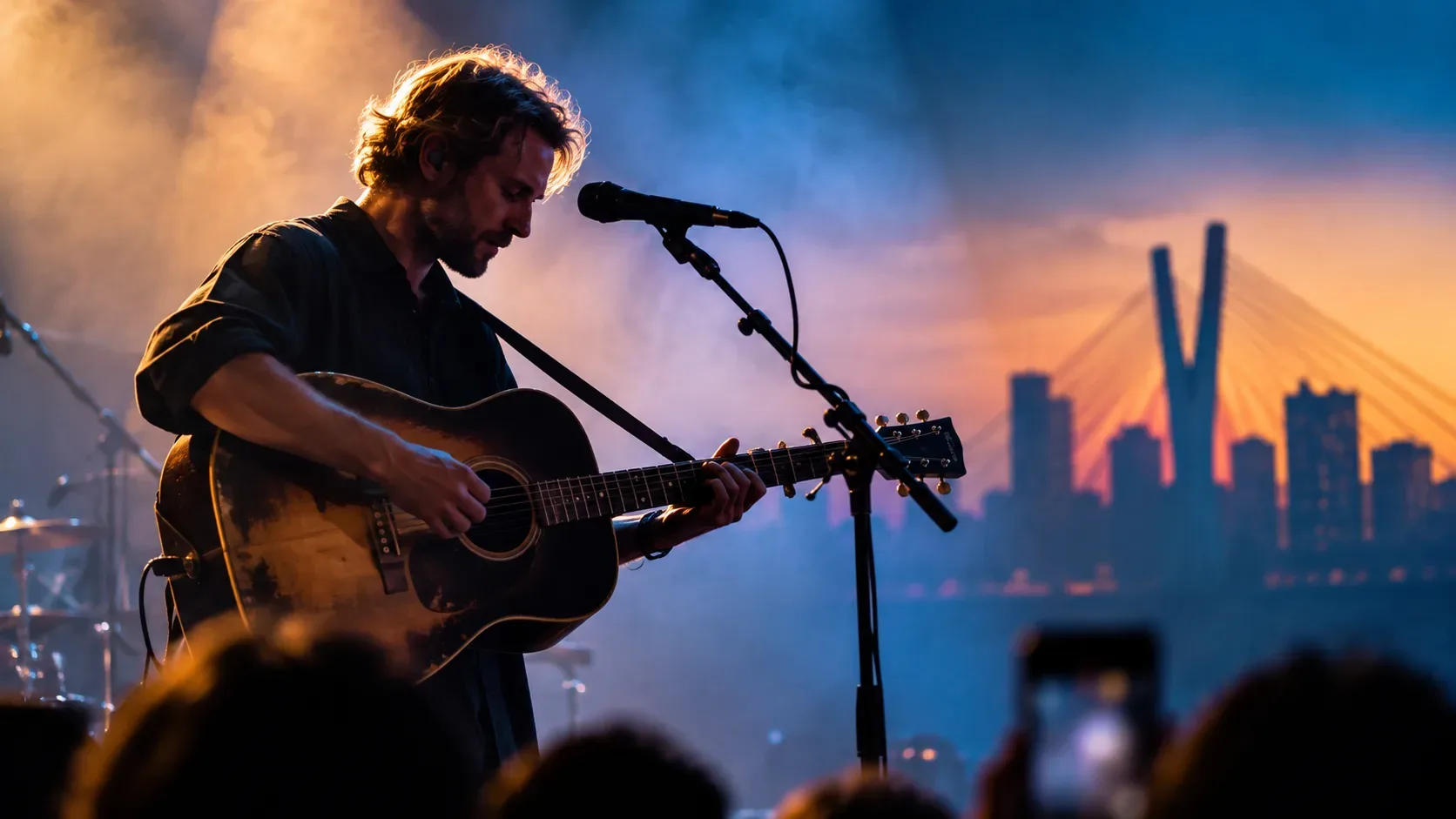 Cantor barbudo toca violão diante de um microfone em palco com luzes azul e dourada, público em primeiro plano e skyline urbano ao fundo.