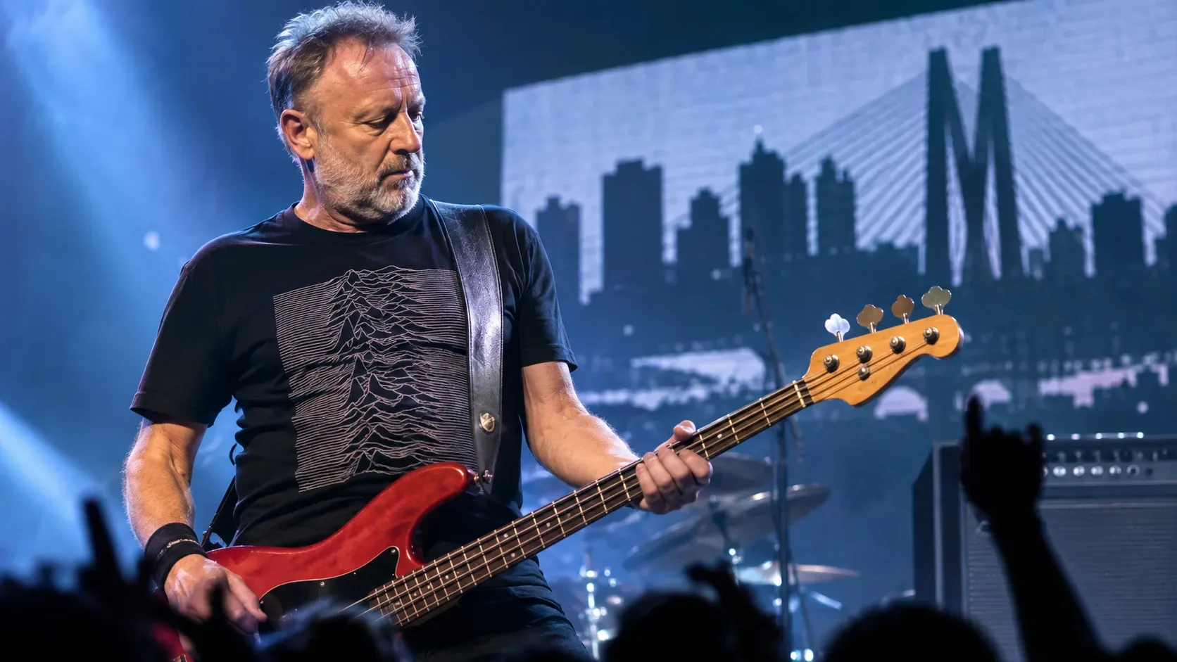 Baixista no palco com baixo vermelho, luzes azuis de show e silhuetas do público diante de cenário urbano de São Paulo.