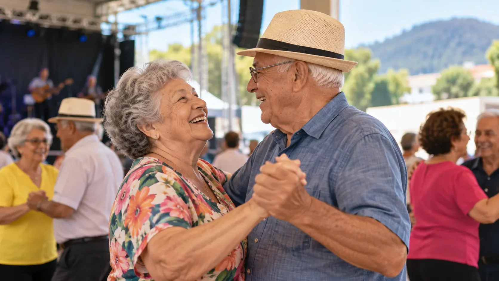 Casal de idosos sorrindo e dançando em um encontro festivo, com outros participantes e um palco ao fundo.