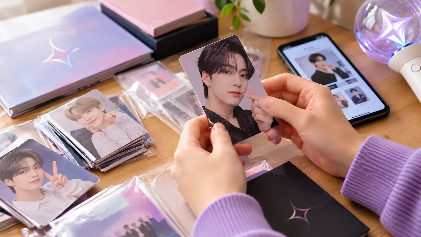 Mãos seguram um photocard de idol sobre mesa com álbuns, sleeves, outros cards, celular e light stick.