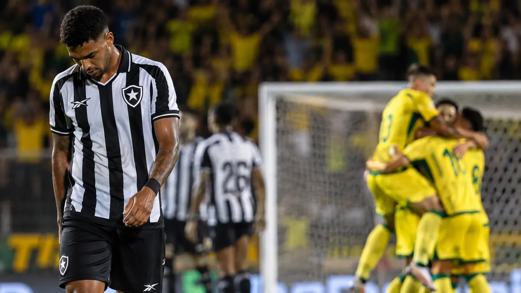 Jogador alvinegro de cabeça baixa em primeiro plano enquanto adversários de amarelo e verde comemoram perto do gol em um estádio à noite.