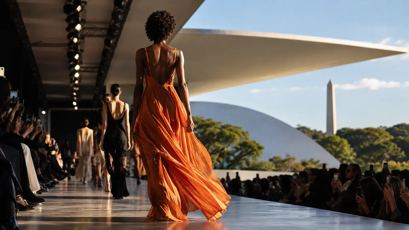 Modelo caminha por uma passarela de moda ao ar livre diante de arquitetura modernista e público no Parque Ibirapuera.