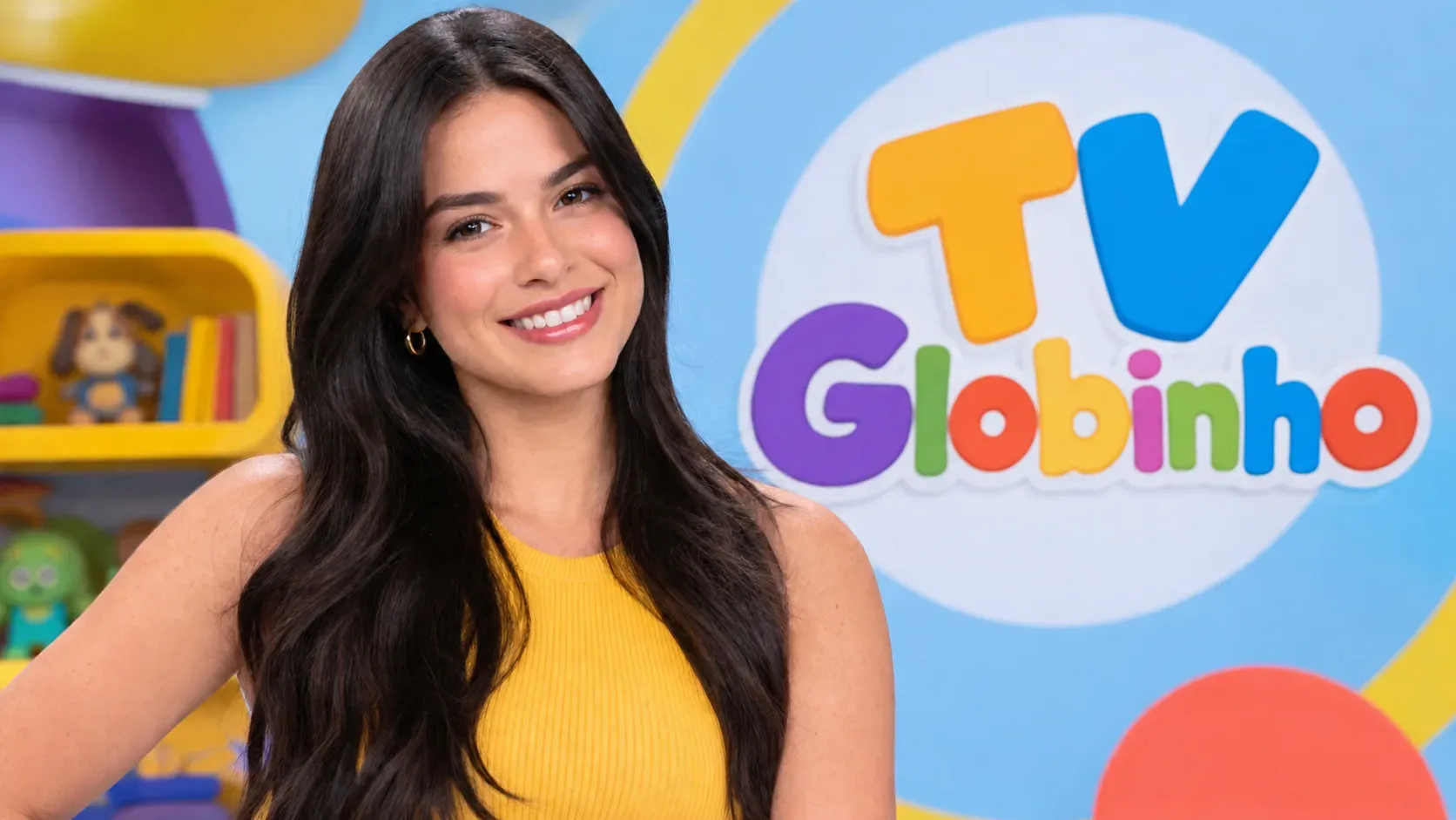 Jovem de cabelos longos escuros e blusa amarela em cenário infantil colorido, com identidade visual da TV Globinho ao fundo.