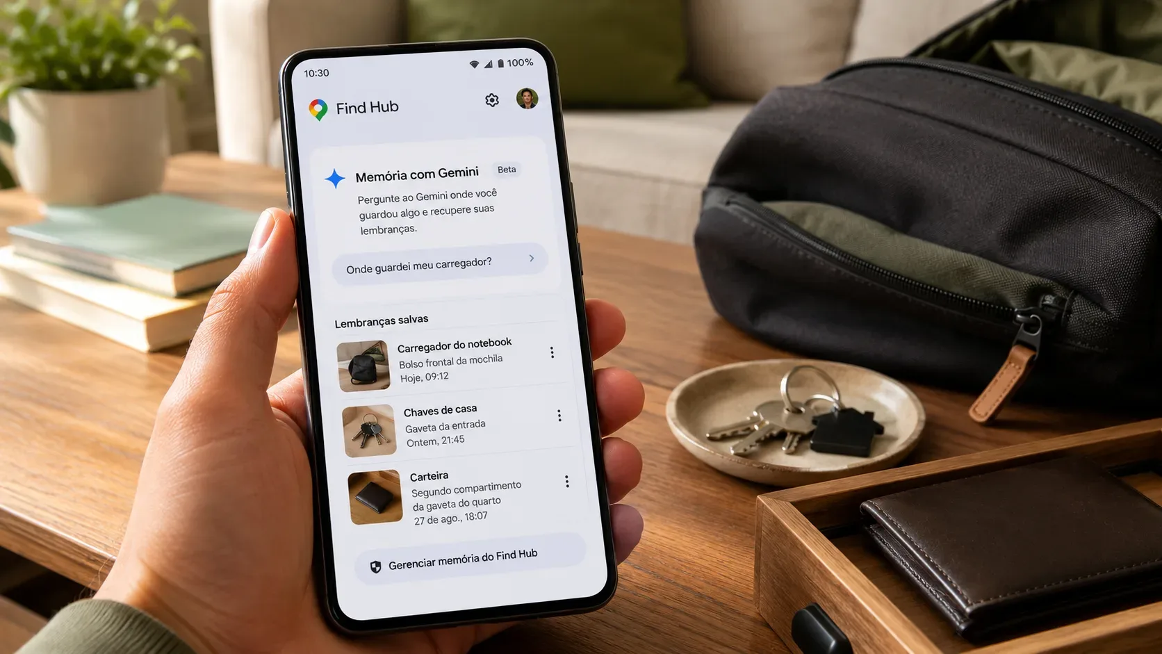 Mão segura um celular Android com a tela do Find Hub aberta, mostrando lembranças salvas de carregador, chaves e carteira sobre uma mesa com mochila e objetos pessoais.